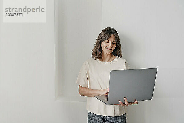 Smiling businesswoman working on laptop in front of wall