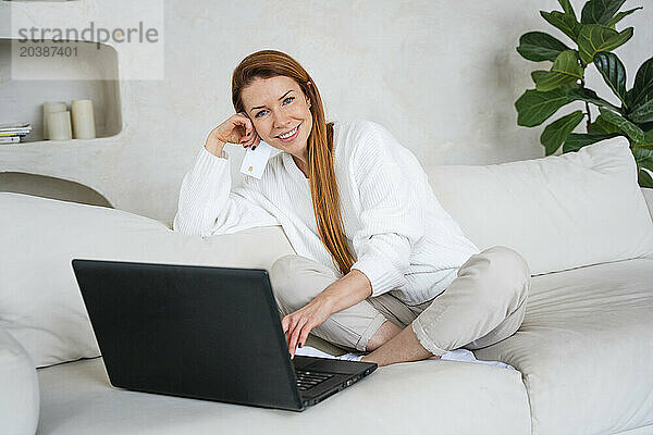 Smiling woman holding credit card shopping online through laptop sitting on sofa at home