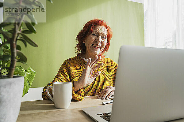 Happy senior woman gesturing on video call through laptop at home