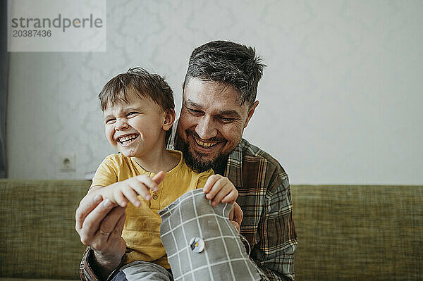 Happy father teaching son to sew button on gray colored cloth at home