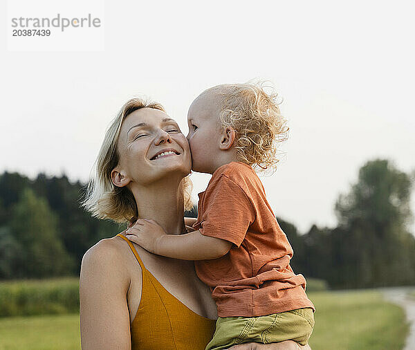 Boy kissing happy mother in meadow