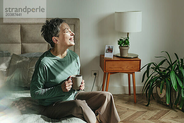 Smiling woman holding coffee mug sitting on bed at home