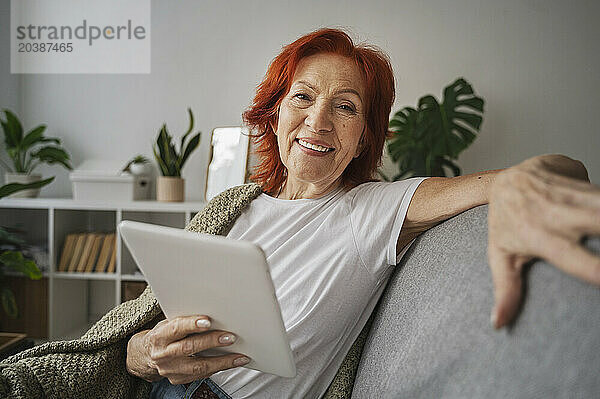 Smiling redhead senior woman with digital tablet sitting on sofa at home