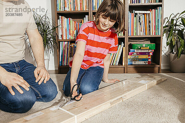 Girl opening plank package with scissors by father kneeling in living room