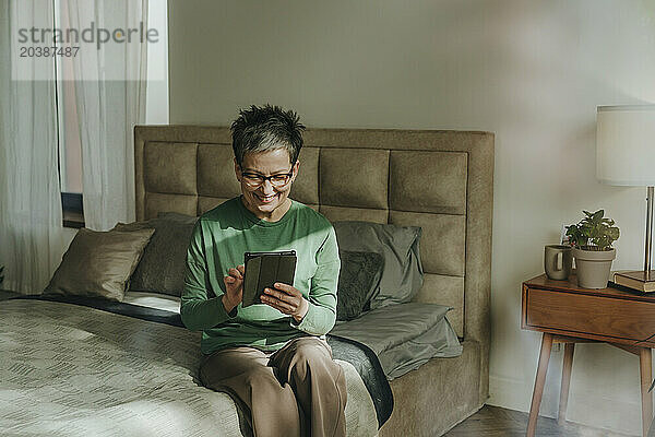 Smiling woman using tablet PC sitting on bed at home