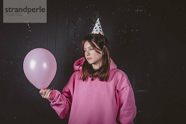 Thoughtful birthday girl in party hat holding pink balloon in front of black background