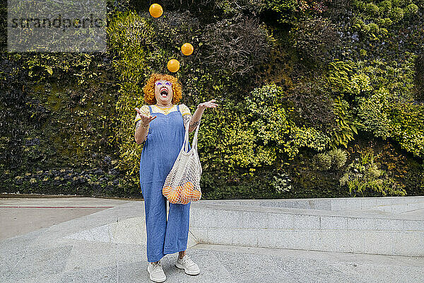 Cheerful woman throwing oranges in air standing on footpath