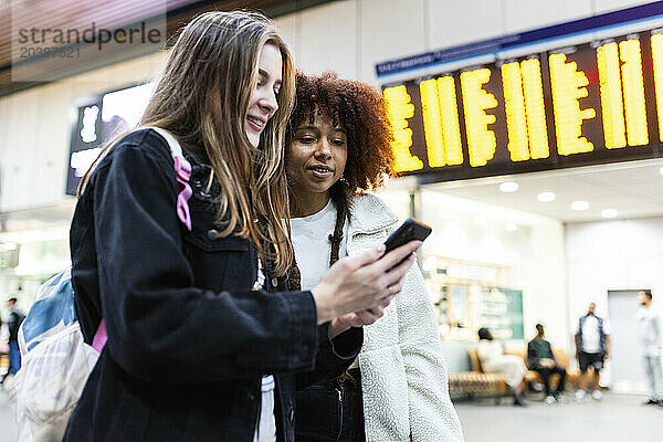 Smiling woman with friend using smart phone at railroad station