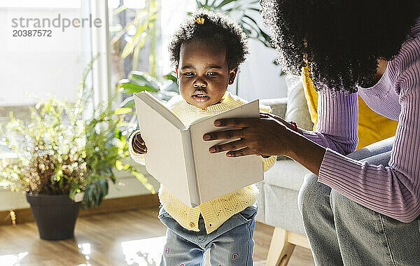 Cute baby girl with mother reading book in living room at home