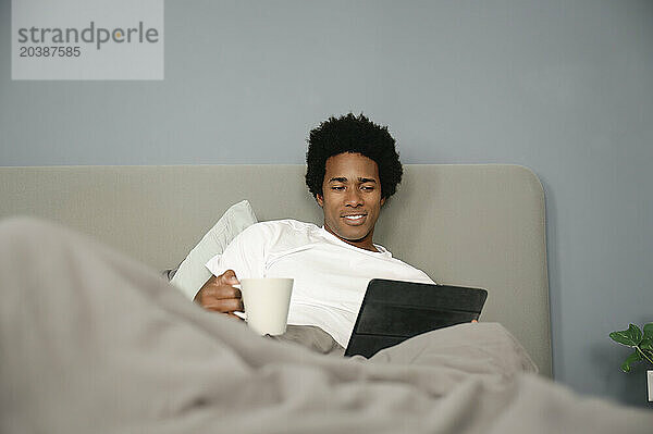 Man using tablet PC relaxing on bed with coffee cup in bedroom at home