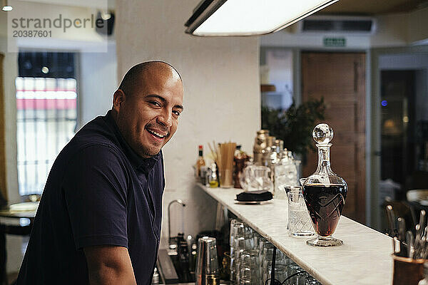 Smiling bartender by alcohol bottle at bar counter in restaurant