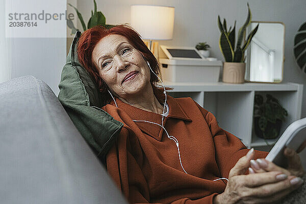 Relaxed redhead senior woman sitting with smart phone and headphones in living room