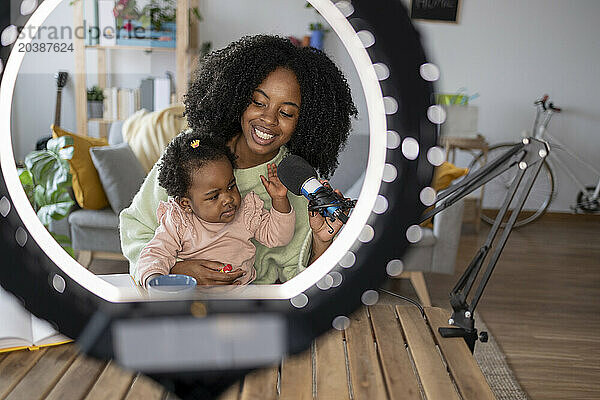 Happy mother and daughter with microphone in front of illuminated ring light at home