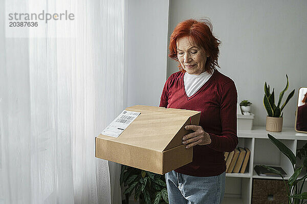 Senior woman opening cardboard box standing by white curtain at home