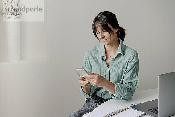 Businesswoman using smart phone sitting near table at home