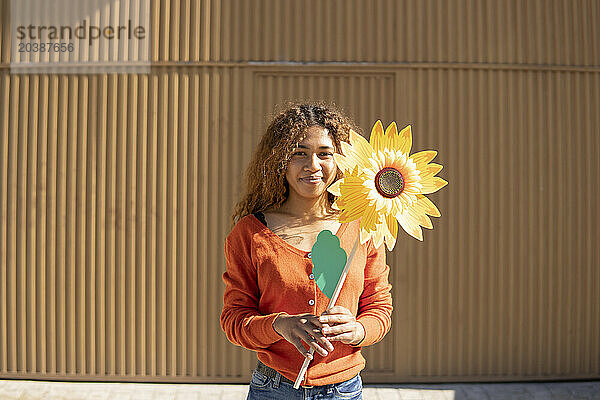 Smiling woman holding artificial sunflower in front of metal wall
