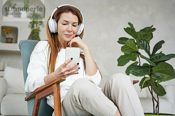 Smiling woman listening music through headphones and using smart phone at home