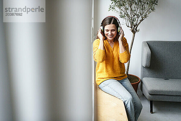 Smiling mature woman listening to music through wireless headphones sitting on window sill at home