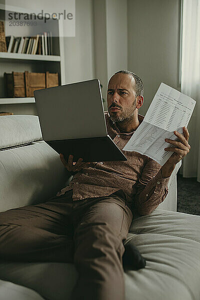 Serious freelancer holding document and looking at laptop in home office