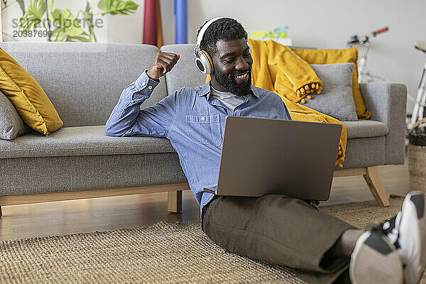 Smiling non-binary person wearing wireless headphones and using laptop in living room at home