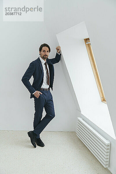 Confident businessman leaning on white wall standing by window at office