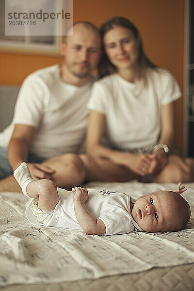 Baby boy lying on bed with parents in background at home