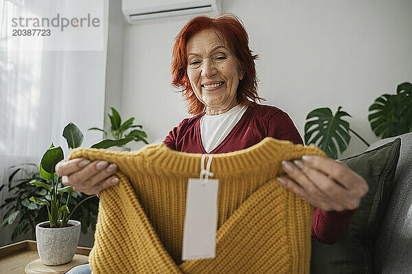 Smiling redhead senior woman examining yellow sweater sitting on sofa at home