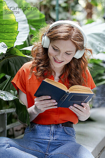 Smiling woman listening to music and reading book sitting on bench