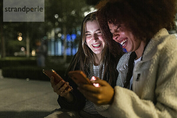 Cheerful young friends using mobile phones at night