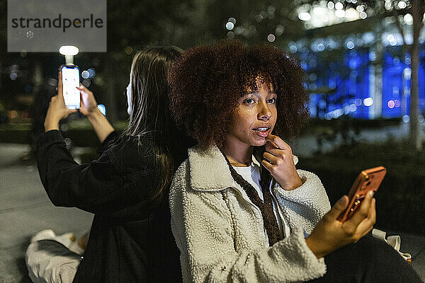 Thoughtful young woman with friend using smart phone at night