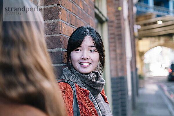 Smiling young woman talking to sister near brick wall