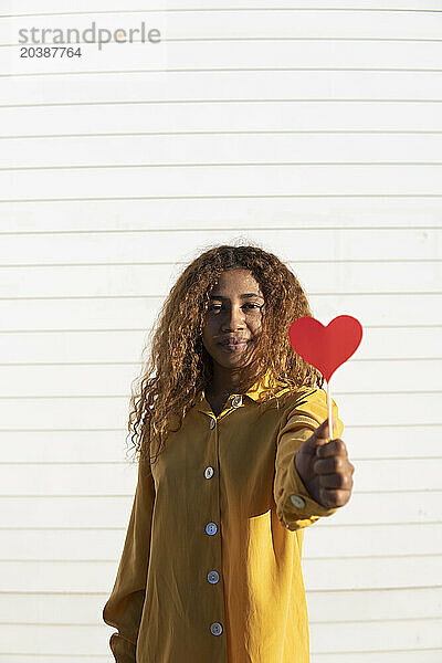 Young curly haired woman giving red heart shape prop in front of white shutter
