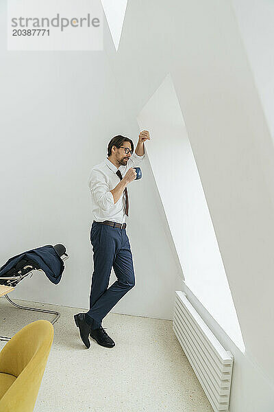 Businessman standing with coffee cup leaning on wall looking out through window at office