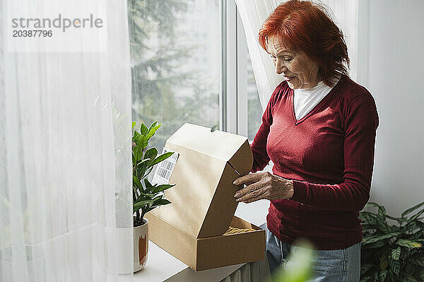 Excited senior woman opening cardboard box standing by window at home