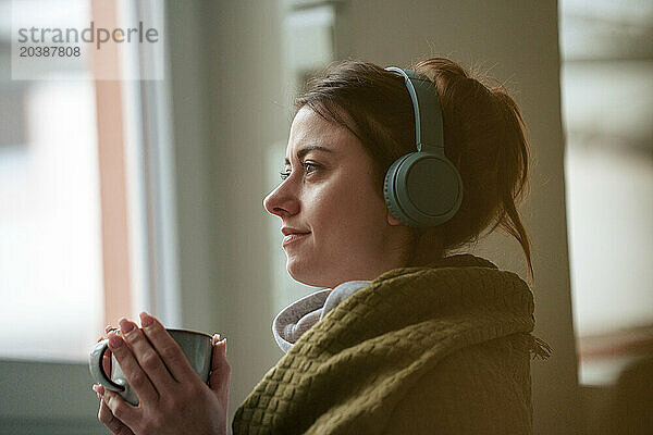 Thoughtful woman listening to music through wireless headphones wrapped in blanket holding coffee cup at home