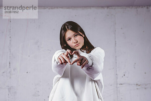 Girl in white hoodie showing heart gesture in front of gray cement background