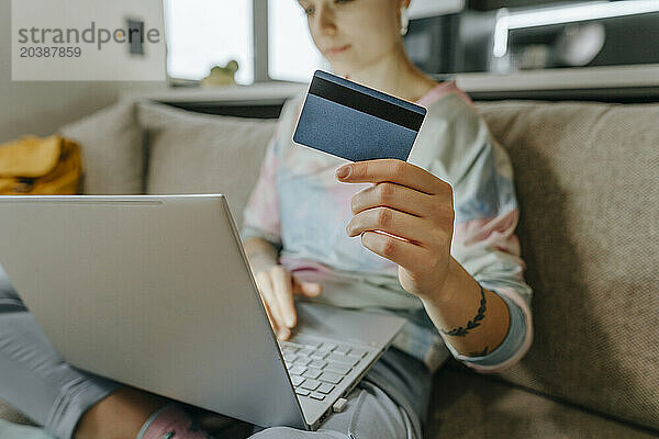 Woman showing credit card sitting with laptop on sofa at home