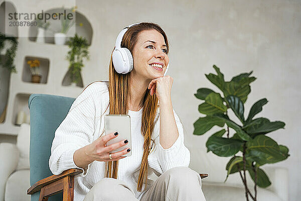 Happy woman listening music through headphones holding smart phone at home