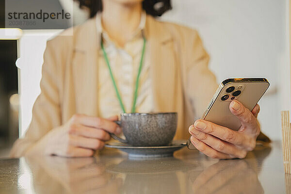 Businesswoman using smart phone sitting with coffee cup at table in cafe