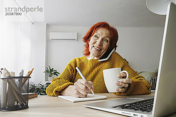 Smiling businesswoman talking on mobile phone sitting at table writing in notepad