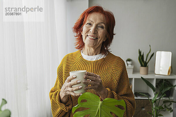 Smiling redhead senior woman standing with tea cup at home