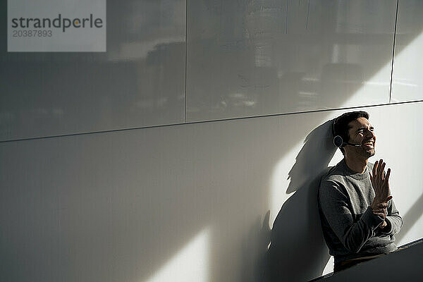 Cheerful businessman talking on headset and sitting near wall in office