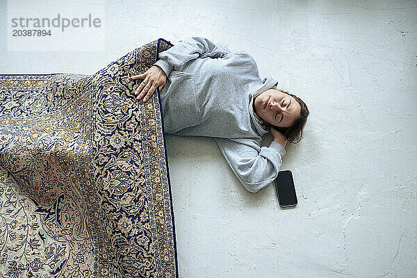 Tired woman sleeping by smart phone on floor under carpet in living room