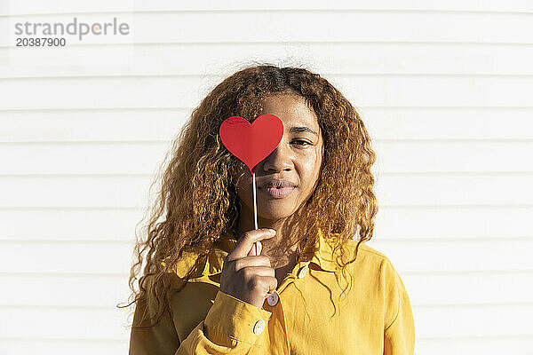 Curly haired woman holding red heart shape prop in front of white shutter