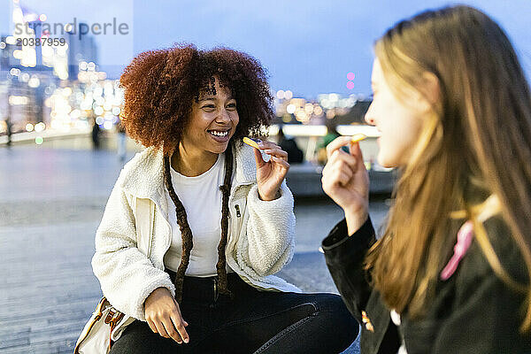 Smiling friends eating french fries in city at dusk