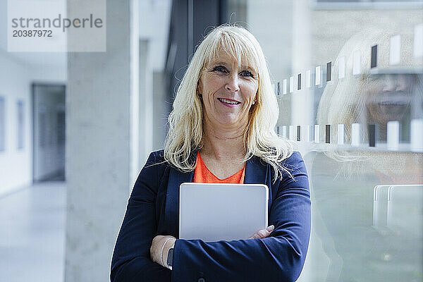 Smiling businesswoman standing with laptop near glass in office corridor