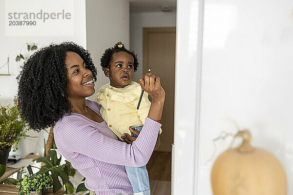 Smiling young single mother carrying daughter writing on refrigerator in kitchen at home