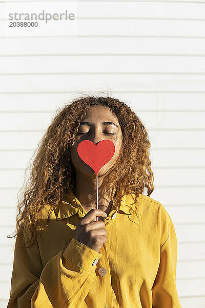 Curly haired woman with eyes closed holding red heart shape prop in front of white shutter