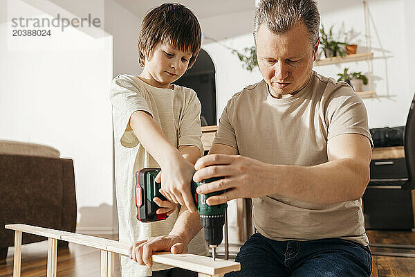 Father assisting son in using power tool for assembling wooden furniture