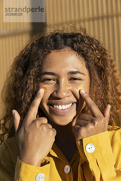 Happy beautiful young curly haired woman pointing at yellow heart shape stickers on cheeks